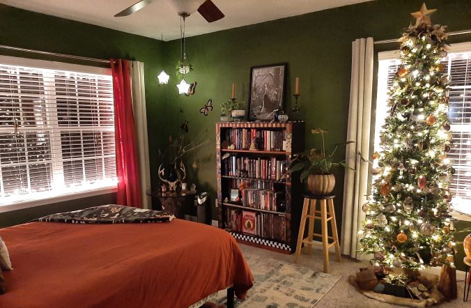 Photo of a swampy green bedroom with Ghibli vibes decorated for Yule/Christmas. The orange and cream curtains are open to reveal windows are framed with white lights. There is a queen bed covered in a dark orange duvet, star lanterns hanging in one corner, and in another, a pencil tree decorated in wildlife and mushrooms with a deerskin tree skirt, feathered topper, and feather ornaments. There is also a bookshelf covered in books and treasures and a figural stump table with a glass topper that looks like an altar but did not originally intend to. :D Beside the tree, sharing the window, is a large Summer Glory philodendron.