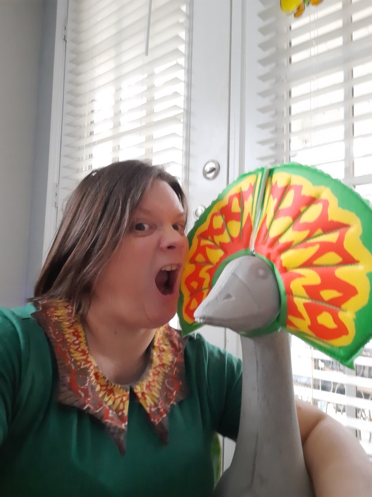 Selfie of me (a middle aged white woman with  a short auburn bob and dark eyes) and Gucifer. He is wearing his dilophosaurus fin and I am wearing my green dilophosaurus dress. I am facing Gucifer with my mouth open as if to bite him.